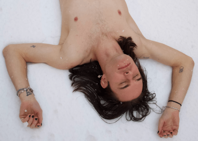 An overhead shot of a shirtless person with long dark hair lying on their back in the snow. Their eyes are closed, and their arms are bent upward at the elbows with palms facing up. Several small tattoos are visible on their torso and arms, and they are wearing bracelets on both wrists and dark nail polish.