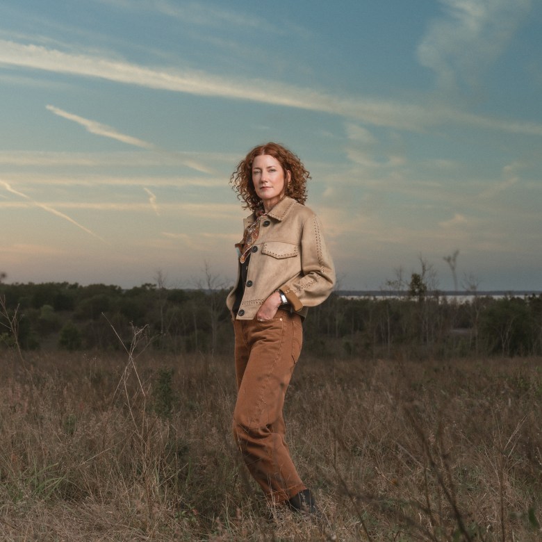 A portrait of a person with curly auburn hair standing in a dry, grassy field at dusk. They are wearing a tan suede jacket over a dark top with a patterned scarf and brown trousers, posing with their hands in their pockets. The background features a vast horizon under a pale blue sky with soft, streaking clouds and a distant line of trees.