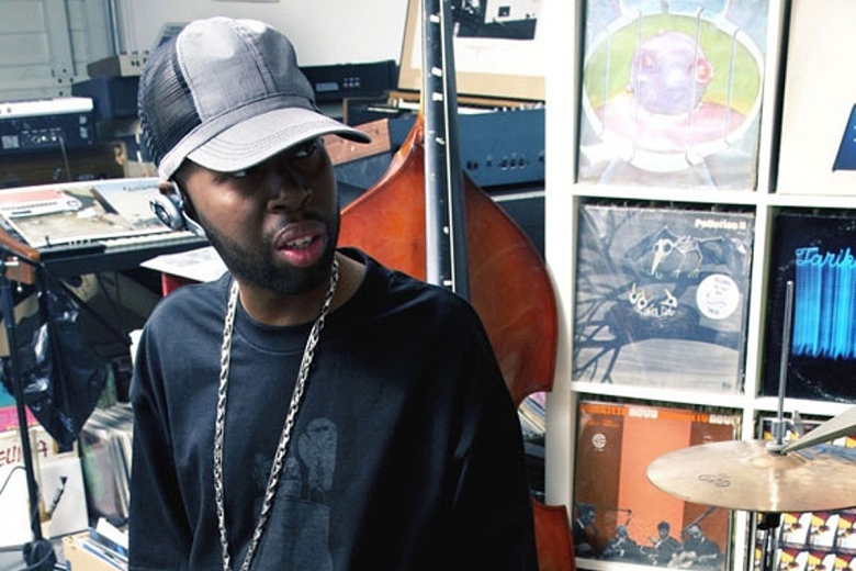 A candid indoor shot of a person with a beard wearing a grey-and-black baseball cap, a black long-sleeved shirt, and a silver chain. They are looking to the side with a concentrated expression. The background is filled with musical equipment and media, including a large upright bass, a drum cymbal in the foreground, and shelves packed with vinyl records and electronics.