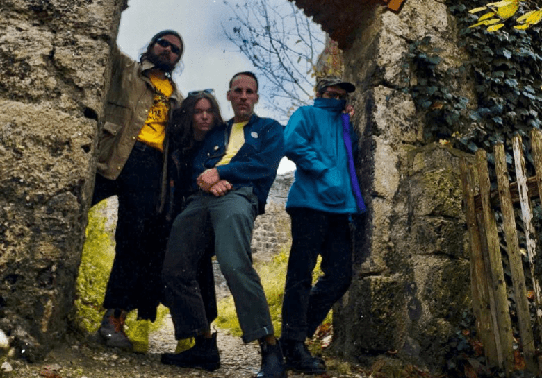 Four people stand together framed within a stone archway of an overgrown, ivy-covered ruin. On the left, a person in a tan jacket and yellow shirt leans against the stone. In the center, two people stand close together—one in a dark blue jacket and the other partially obscured behind them. On the right, a person in a bright blue-and-purple jacket and a camouflage cap looks toward the camera. A weathered wooden fence is visible on the far right against the mossy stone walls.