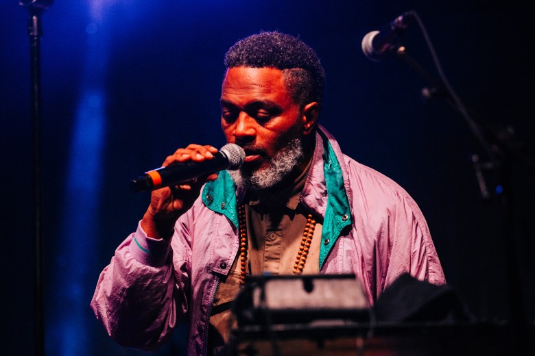 A close-up, dramatic live performance shot of a person with a grey-flecked beard and short hair singing into a microphone. They are wearing a light pink windbreaker with a teal collar over a brown button-down shirt and a long beaded necklace. The scene is bathed in atmospheric blue and red stage lighting, with a second microphone stand visible in the blurred background.