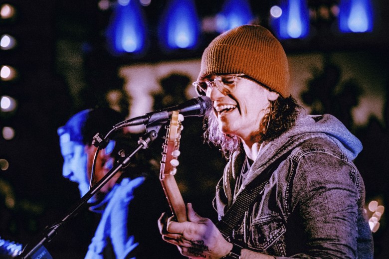 A close-up, live performance shot of a musician singing into a microphone while playing an acoustic guitar. They are smiling and wearing a brown beanie, clear-framed glasses, and a grey denim jacket over a hoodie. The background is softly blurred with blue and white bokeh lights, and the profile of another performer is visible in a blue hue on the left.