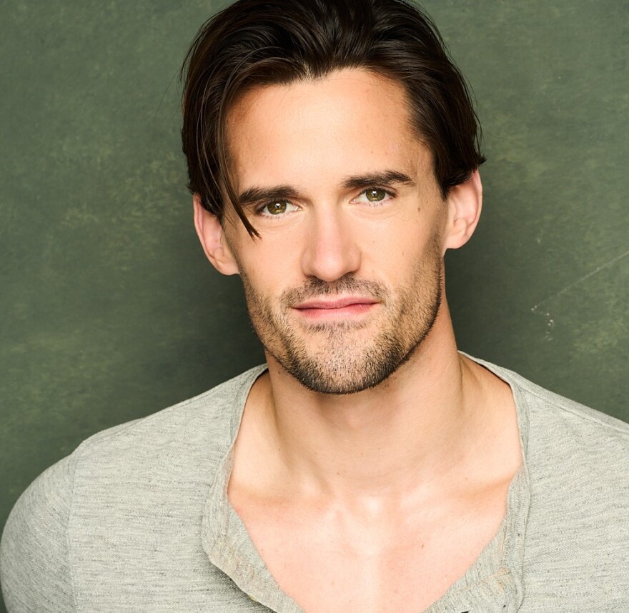 A young man with dark hair and brown eyes with a scruffy beard and mustache, dressed in a grey Henley, looks at the camera