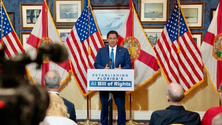 Gov. Ron DeSantis speaking at a podium labeled "Establishing Florida's AI Bill of Rights" with American Flags behind him and a tv camera and audience members in the foreground.