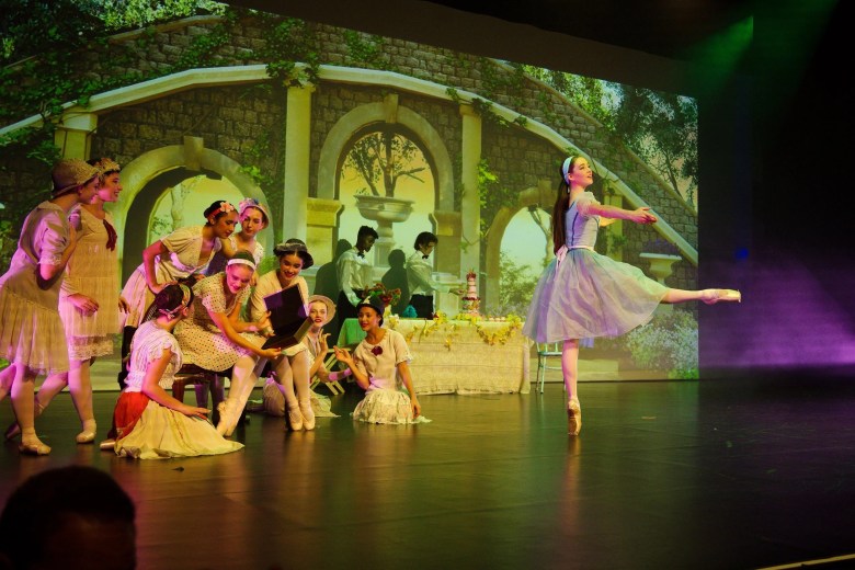 Ballet dancers perform on stage in front of a backdrop depicting stone arches and a garden scene. A group of dancers in pastel costumes gather around a box at center stage, while another dancer in a blue dress balances on pointe with one leg extended gracefully to the side. A table decorated with flowers and a cake is set in the background, creating a festive atmosphere.