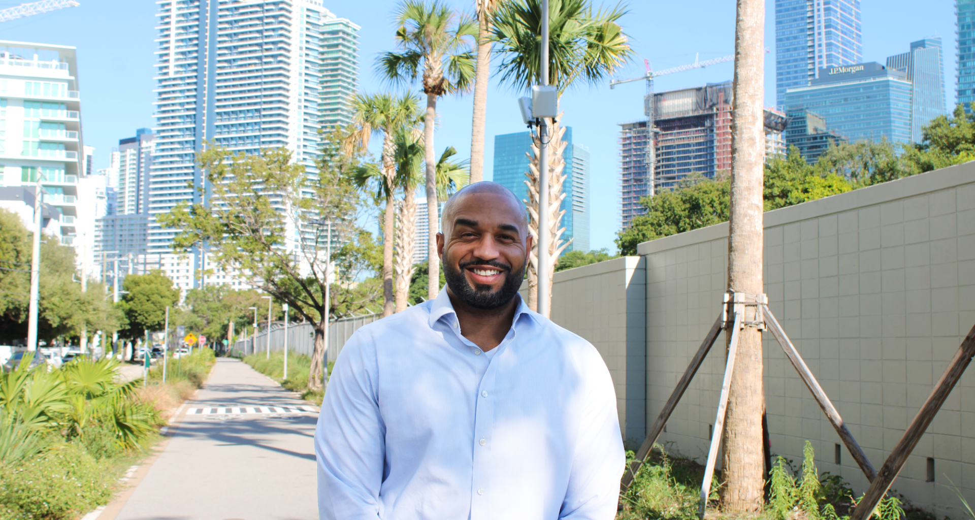 photo of a bearded Black man in a button-down shirt posing in a pedestrian pathway in with skyscrapers in the distance