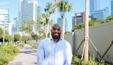 photo of a bearded Black man in a button-down shirt posing in a pedestrian pathway in with skyscrapers in the distance