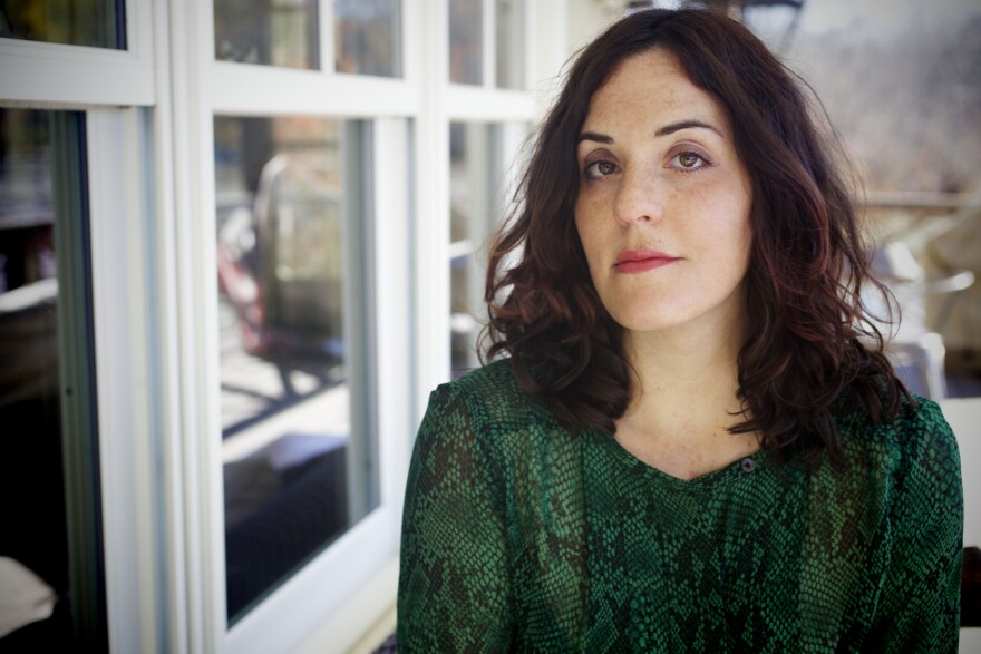 A woman with shoulder length dark brown hair, wearing a green top, looks at camera.