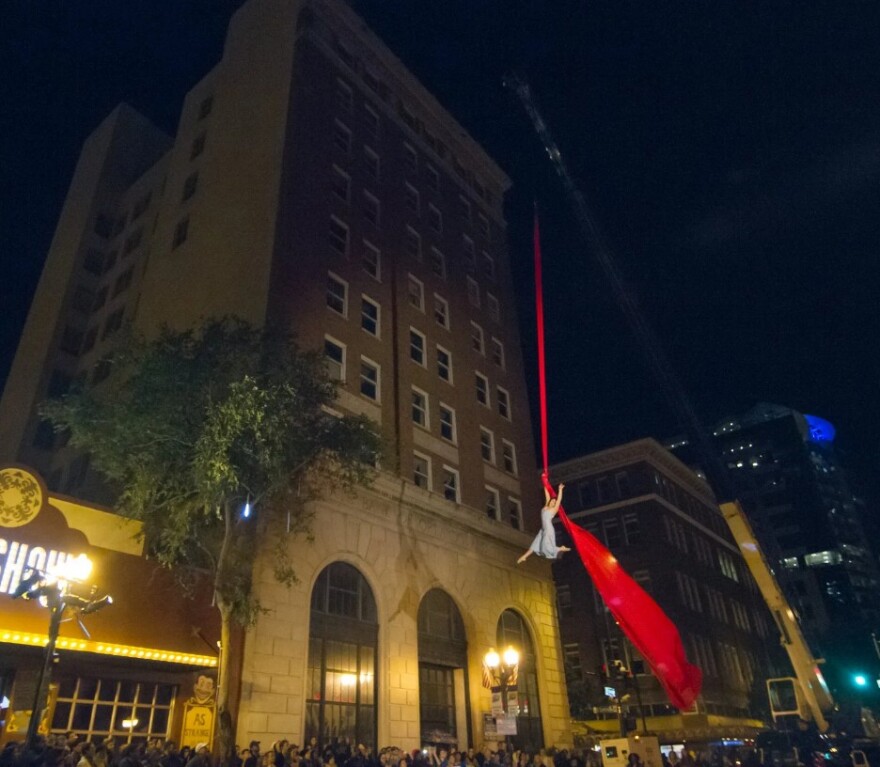 A Cirque du Soleil aerial artist performs while hanging from a crane near the top of a downtown building during Immerse 2025.