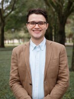 Man with dark hair and glasses wearing a brown sport jacket and light shirt smiling into the camera with trees and grass in the background