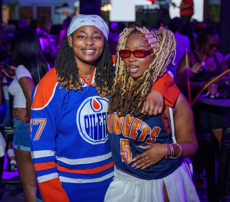 A pair of partygoers at a Her Way event. Two people pose arm-in-arm at a dimly lit party illuminated by purple club lights. The person on the left wears a white bandana and a blue Edmonton Oilers hockey jersey. The person on the right wears red rectangular sunglasses, a blue Denver Nuggets basketball jersey, and a white pleated skirt. Their blonde locs are styled with colorful hair clips. Other patrons and part of a hookah pipe are visible in the blurred background.