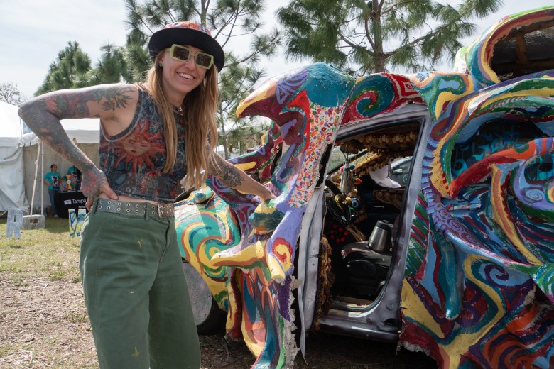 A smiling woman with arm tattoos standing next to a brightly colored, highly textured art car at Gasparilla Festival of the Arts.