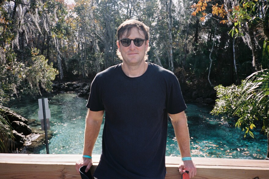 A man in a black t-shirt wearing sunglasses outside on a bridge above some water