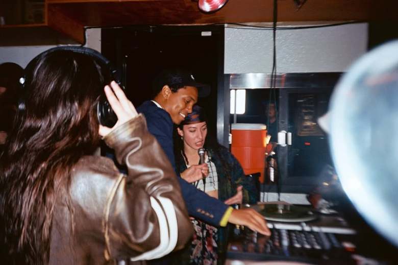 Tampa artist Taína Enserio holding a microphone and talking in a DJ booth next to a performer at a crowded indoor event.