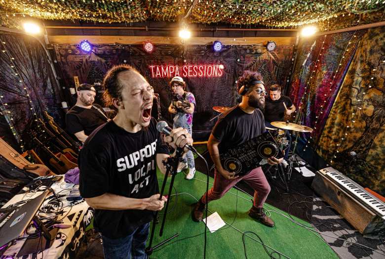 Tampa songwriter and poet Dennis Amadeus of Wolfgang Amadeus screaming into a microphone during a live performance at Tampa Sessions, wearing a black "Support Local Art" T-shirt with a full band on a green turf stage.