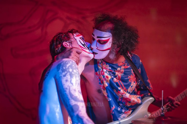 Members of the Alabama-based surf rock band Daikaiju perform a theatrical, high-energy set in Tampa. One performer in a floral shirt plays guitar while another with blue body paint leans in close; both wear the band's signature kabuki-style masks under red stage lights.