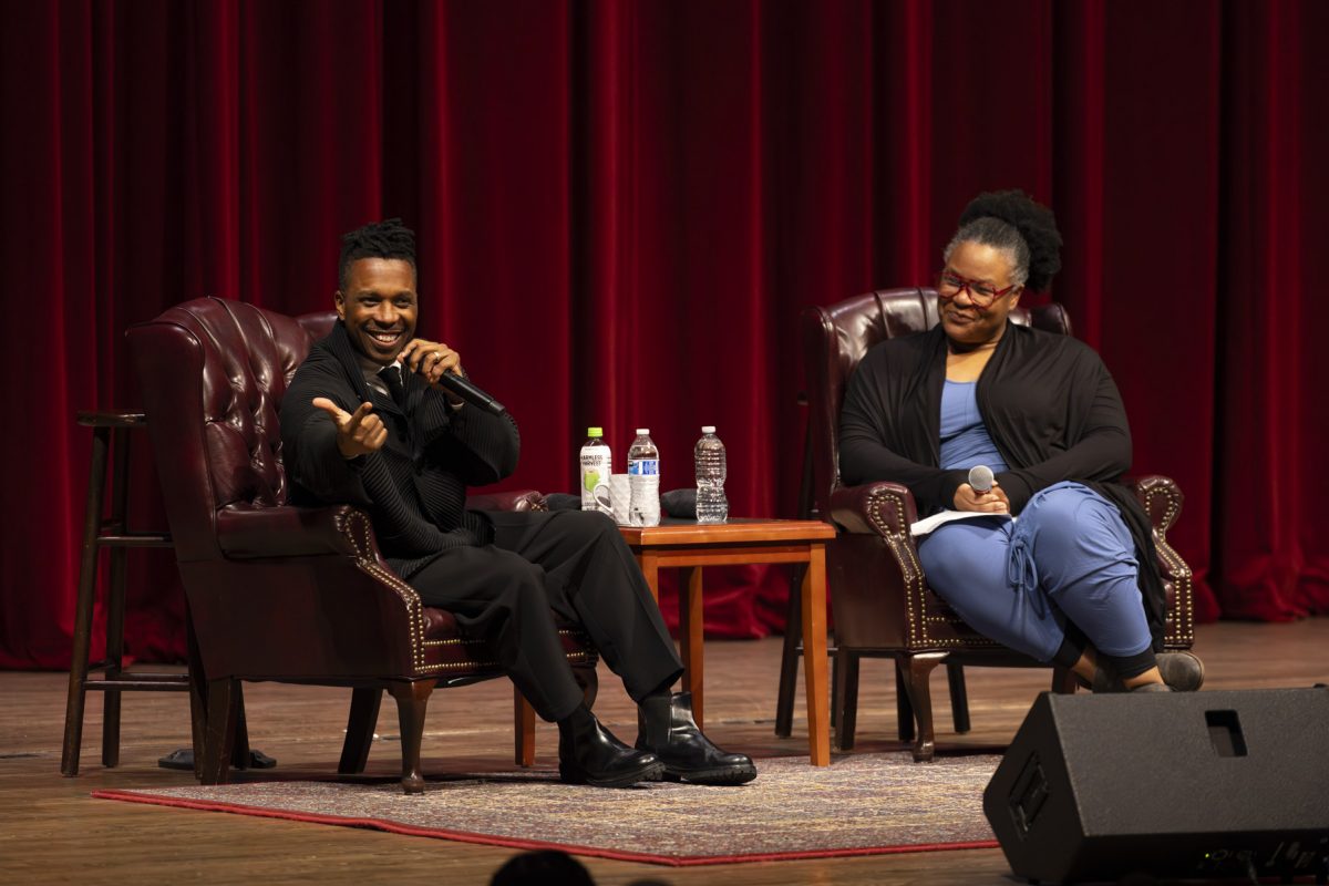 Two people sitting on chairs on a stage, one of them speaking expresisvely into a microphone