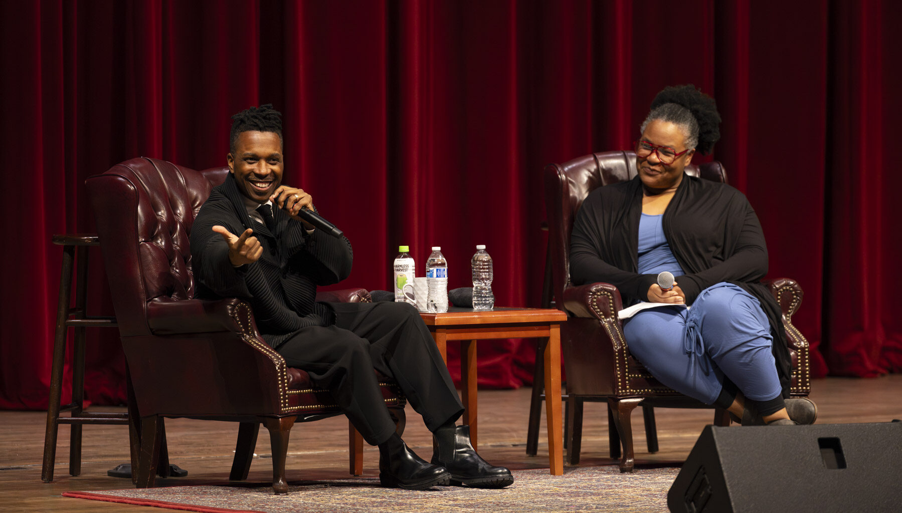 Two people sitting on chairs on a stage, one of them speaking expresisvely into a microphone