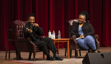Two people sitting on chairs on a stage, one of them speaking expresisvely into a microphone
