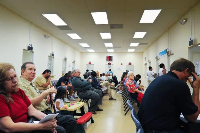People seated in a busy waiting room, some reading, others using phones, with numbered kiosks visible in the background
