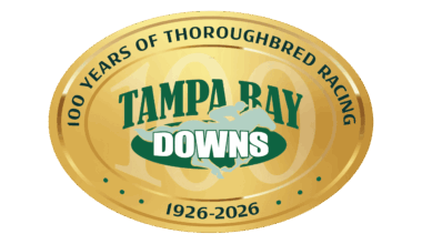 The 100 years anniversary logo for Tampa Bay Downs. The logo is a golden, oval emblem, with thin outlines surrounding the circumference of the emblem that make the vector look like shining gold. Dark green curving text along the outside of the logo reads, “100 Years of Thoroughbred Racing, 1926 - 2026.” In the middle of the logo, Tampa Bay Downs standard brand logo (consisting of a galloping racehorse and jockey, overlapping the name “Tampa Bay Downs” in dark and light green) is centered overtop a large “100,” which is partially transparent and fades into the gold of the logo.