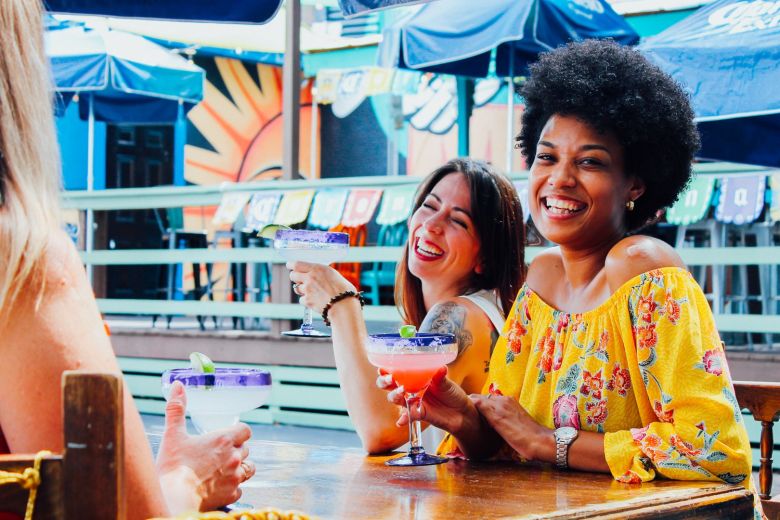 A group of friends laughing and enjoying margaritas together at Nueva Cantina's colorful outdoor patio bar.