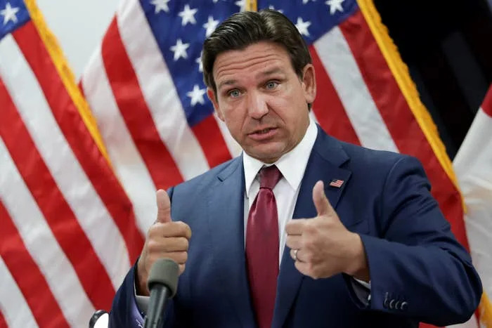 Person in a suit gives thumbs up at a podium in front of American flags