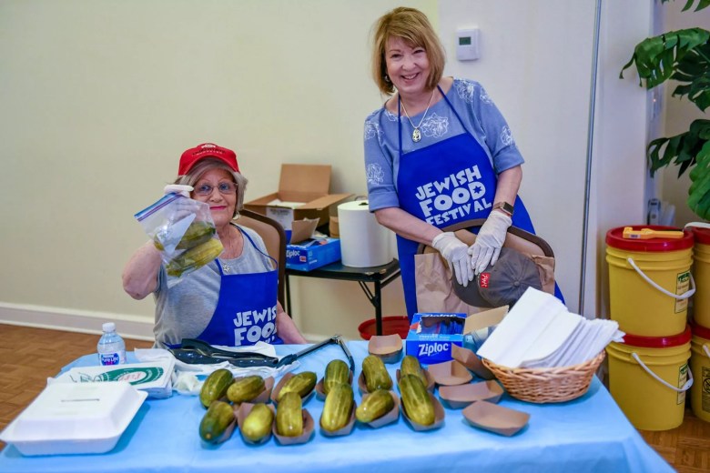 Two women wearing blue aprons reading 'JEWISH FOOD FESTIVAL' smile in front of a table of large pickles. 