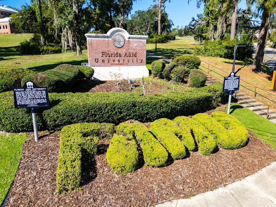 Shrubbery spelling "FAMU" in front of a brick Florida A&M University sign with surrounding greenery and historical markers