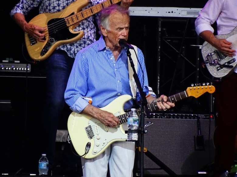 Al Jardine of The Beach Boys, in a blue-and-white striped button-down shirt and white pants stands on a stage, playing a cream-colored electric guitar. He is singing into a microphone attached to a stand in front of him. A black guitar strap is slung over his shoulder. In the background, two other musicians are partially visible, with another electric guitar and a bass guitar. A Fender amplifier is positioned behind the main musician. The stage is set with various music equipment, including speakers and a keyboard stand.