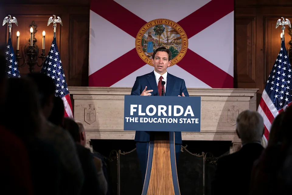 A man speaks at a podium labeled "Florida: The Education State" in front of a Florida flag and two U.S. flags, addressing an audience
