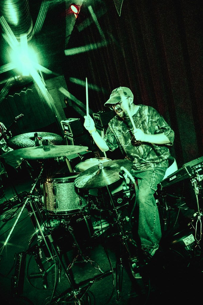 A high-energy, live music photograph of a drummer performing under intense green stage lighting. The drummer, wearing a camouflage t-shirt, glasses, and a baseball cap, is captured mid-motion with drumsticks raised above a silver sparkle drum kit. The scene is filled with dramatic light flares and shadows, creating a gritty, atmospheric feel.