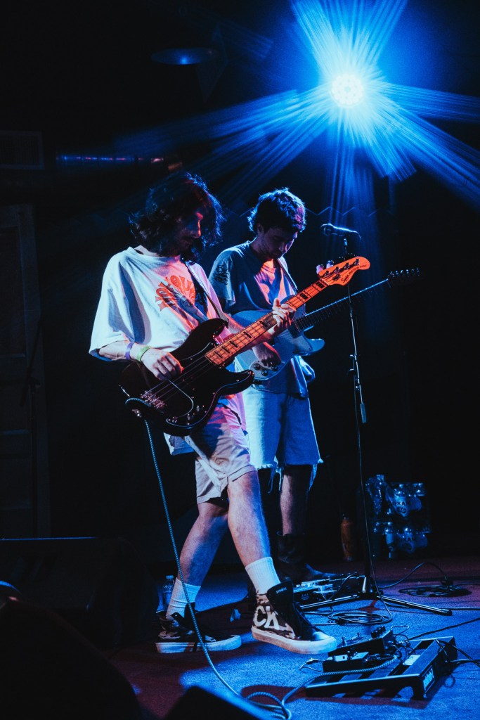 A live performance shot of two musicians on a dark stage illuminated by a bright blue spotlight with a starburst flare. In the foreground, a person with long hair and a white graphic t-shirt plays a black bass guitar. Behind them, another person in a grey shirt plays an electric guitar. Both are focused on their instruments, with various effect pedals and cables visible on the stage floor.