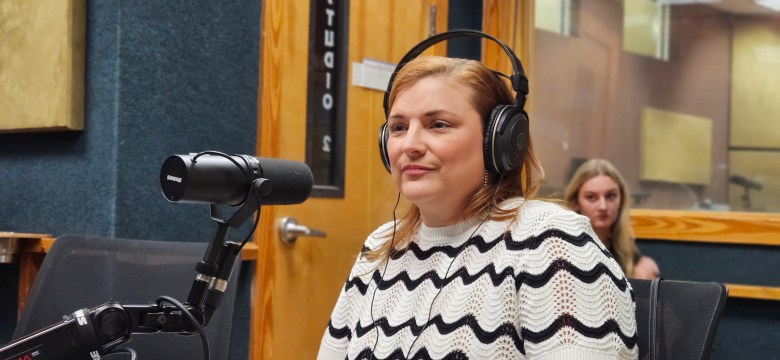 A person with auburn hair wearing large black headphones and a black-and-white wavy-patterned sweater. They are seated in a radio studio, looking slightly off-camera with a neutral expression. A black professional Shure microphone is positioned in the foreground, and a wooden door with "STUDIO 2" marked on it is visible in the background.