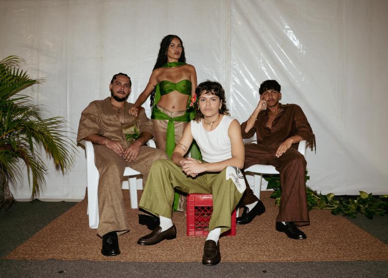 Four people pose for a group portrait in front of a white draped backdrop. One person stands in the center back wearing a green bandeau top and a matching wrap, while three others are seated in front of them: one on the left in a tan textured suit, one in the center foreground on a red plastic crate wearing a white tank top and green trousers, and one on the right in a brown button-down set. They are positioned on a brown rug, and a potted palm tree is visible on the far left.