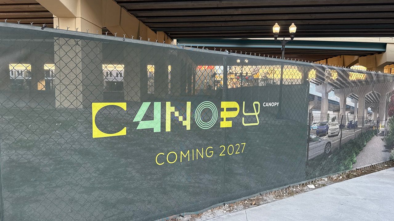 A green signs reads The Canopy Project Coming 2027, mounted to a fence underneath the elevated section of Interstate 4 in downtown Orlando. (Spectrum News/Keith Landry)