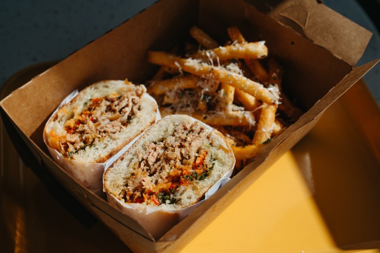 A sliced porchetta sandwich with herb stuffing and crispy skin, served with parmesan fries in a takeout box at Chicken and Pig in Mulberry, FL.