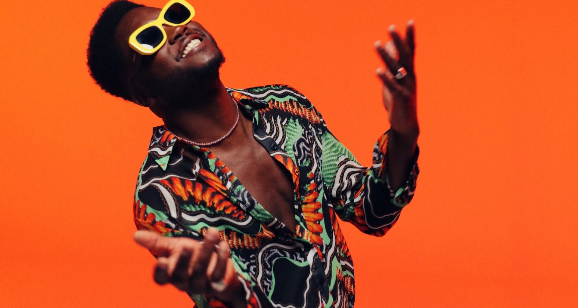 A man wearing yellow glasses and a colorful shirt against an orange background