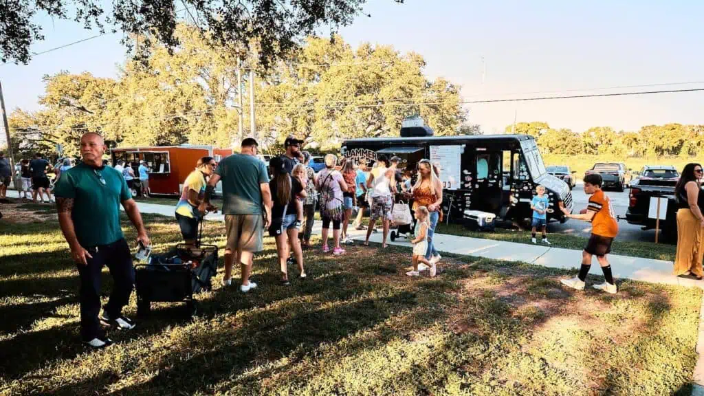 a big line forms near a big food truck that's stationed in parking lot of a community park