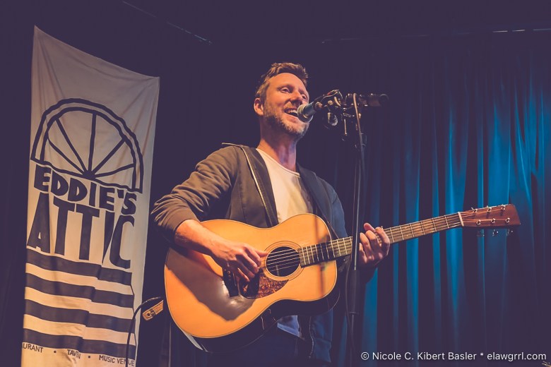 Singer-songwriter Cory Branan performs live on stage at Eddie's Attic. He is smiling while singing and playing an acoustic guitar, positioned in front of a blue curtain and a banner for the music venue.