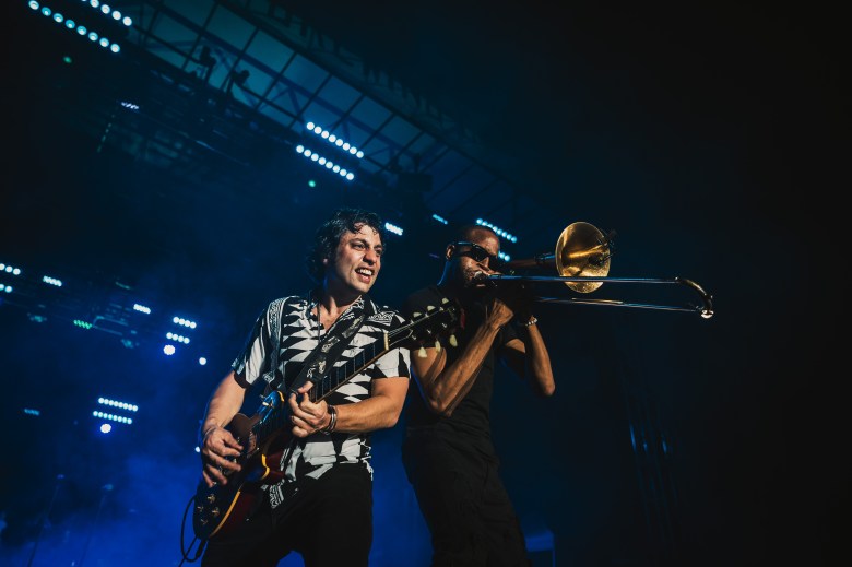 Trombone Shorty (Troy Andrews) performing on trombone alongside guitarist Pete Murano at Curtis Hixon Waterfront Park in downtown Tampa. The musicians are on an outdoor stage at night, illuminated by blue stage lights against a dark sky.