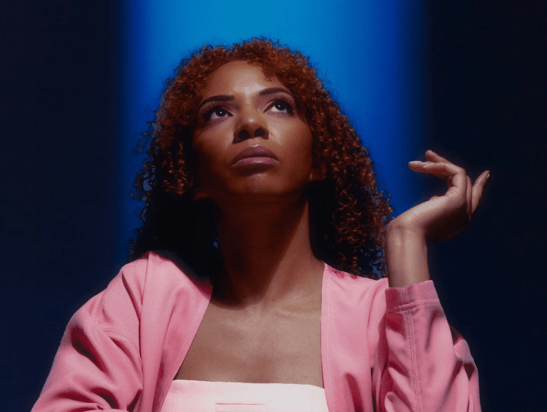 DJ Holographic looking upward against a high-contrast blue and black studio background, featuring her signature red curly hair and a pink jacket.