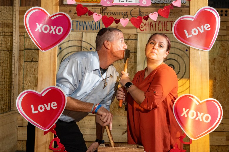 A couple posing playfully with axes inside a Valentine's Day photo frame decorated with red and pink hearts.