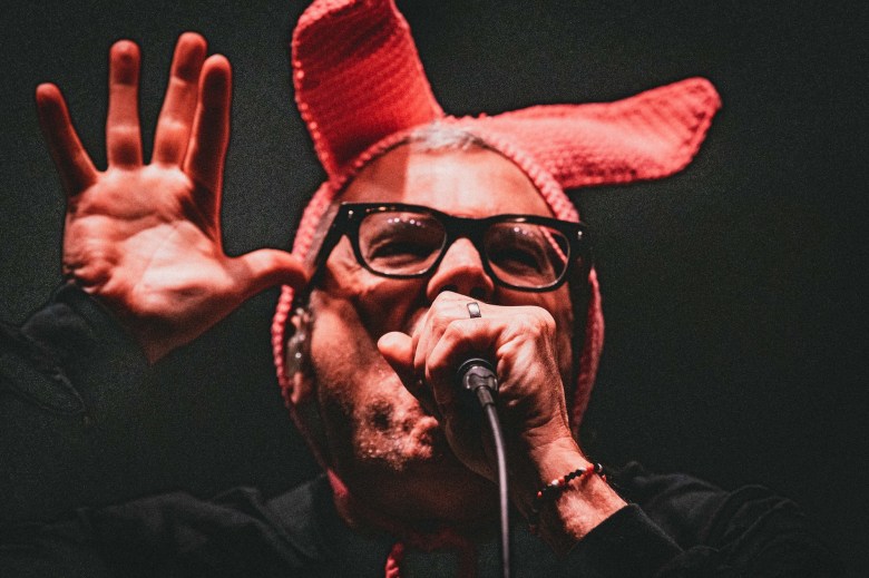 Milo Aukerman of the punk band Descendents performing live on stage in a red knitted bunny ear hat, singing into a microphone with one hand raised against a dark background.