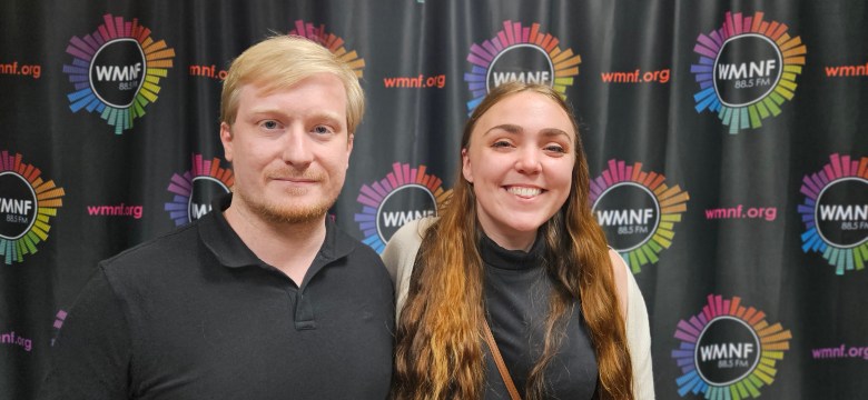 A medium-shot portrait of two people standing together in front of a black backdrop patterned with colorful "WMNF 88.5 FM" logos. The individual on the left has short blonde hair and a light beard, wearing a black polo shirt. The individual on the right has long wavy brown hair and is smiling, wearing a black top and a cream-colored cardigan.