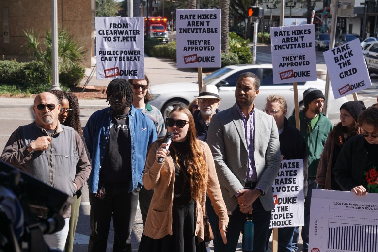 A person with long brown hair stands at a street corner, speaking into a handheld microphone while wearing sunglasses and a tan cardigan. They are surrounded by several other people holding protest signs that read "RATE HIKES AREN'T INEVITABLE," "TAKE BACK THE GRID," and "PUBLIC POWER CAN'T BE BEAT." The background shows a sunny city street with palm trees and a red emergency vehicle in the distance.