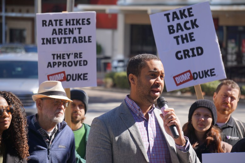 A close-up of a person in a grey blazer and checkered button-down shirt speaking into a microphone. In the background, other participants are visible, including one person in a straw hat and another in a black beanie. Two protest signs are held high behind the speaker, reading "RATE HIKES AREN'T INEVITABLE" and "TAKE BACK THE GRID," both featuring the "DUMP DUKE" logo.