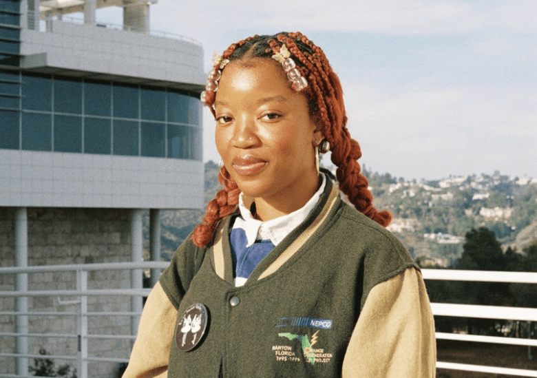 Portrait of Tampa Bay songwriter Fessi K wearing a green and tan letterman-style varsity jacket. She has reddish-brown braided hair adorned with butterfly clips and beads, posing outdoors in front of a modern white building and a hilly landscape.