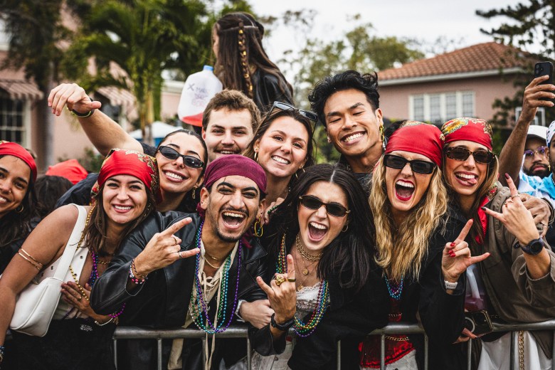 A high-energy group portrait of approximately ten people celebrating at the Gasparilla Pirate Fest. Many are wearing pirate-themed attire, including red bandanas, gold jewelry, sunglasses, and colorful beaded necklaces. They are smiling and shouting toward the camera, with several individuals making "rock on" hand gestures while leaning against a metal crowd-control barricade. The outdoor background features palm trees and a residential building under an overcast sky.
