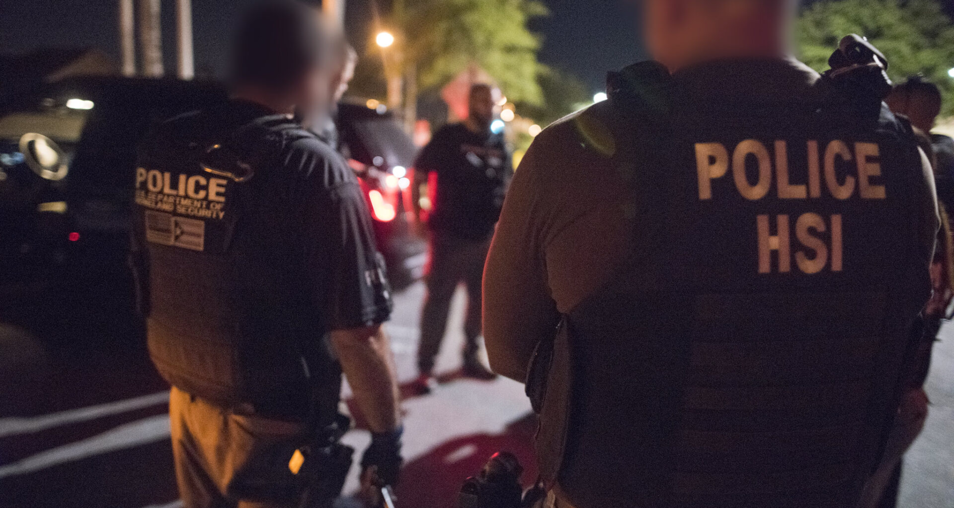 Two Department of Homeland Security agents face away from the camera outdoors at night